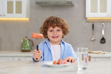 Cute little boy holding fork with sausages at table in kitchen