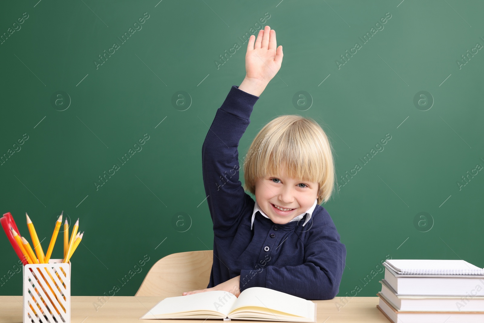 Photo of Happy little school child raising hand while sitting at desk with books near chalkboard