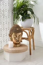 Photo of Golden Buddha head and burning candle on wicker pouf indoors. Interior design