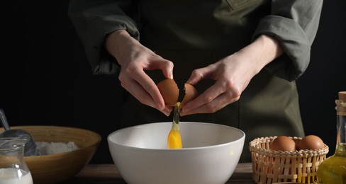 Photo of Making bread. Woman adding egg into dough at wooden table on dark background, closeup