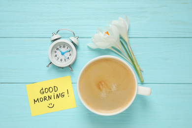 Photo of Delicious coffee, alarm clock, flowers and card with GOOD MORNING wish on light blue wooden table, flat lay
