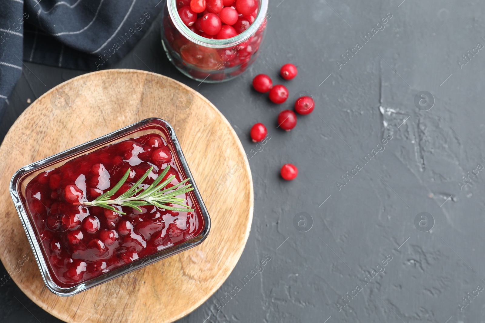 Photo of Tasty cranberry sauce in bowl and glass jar with fresh berries on gray textured table, flat lay. Space for text