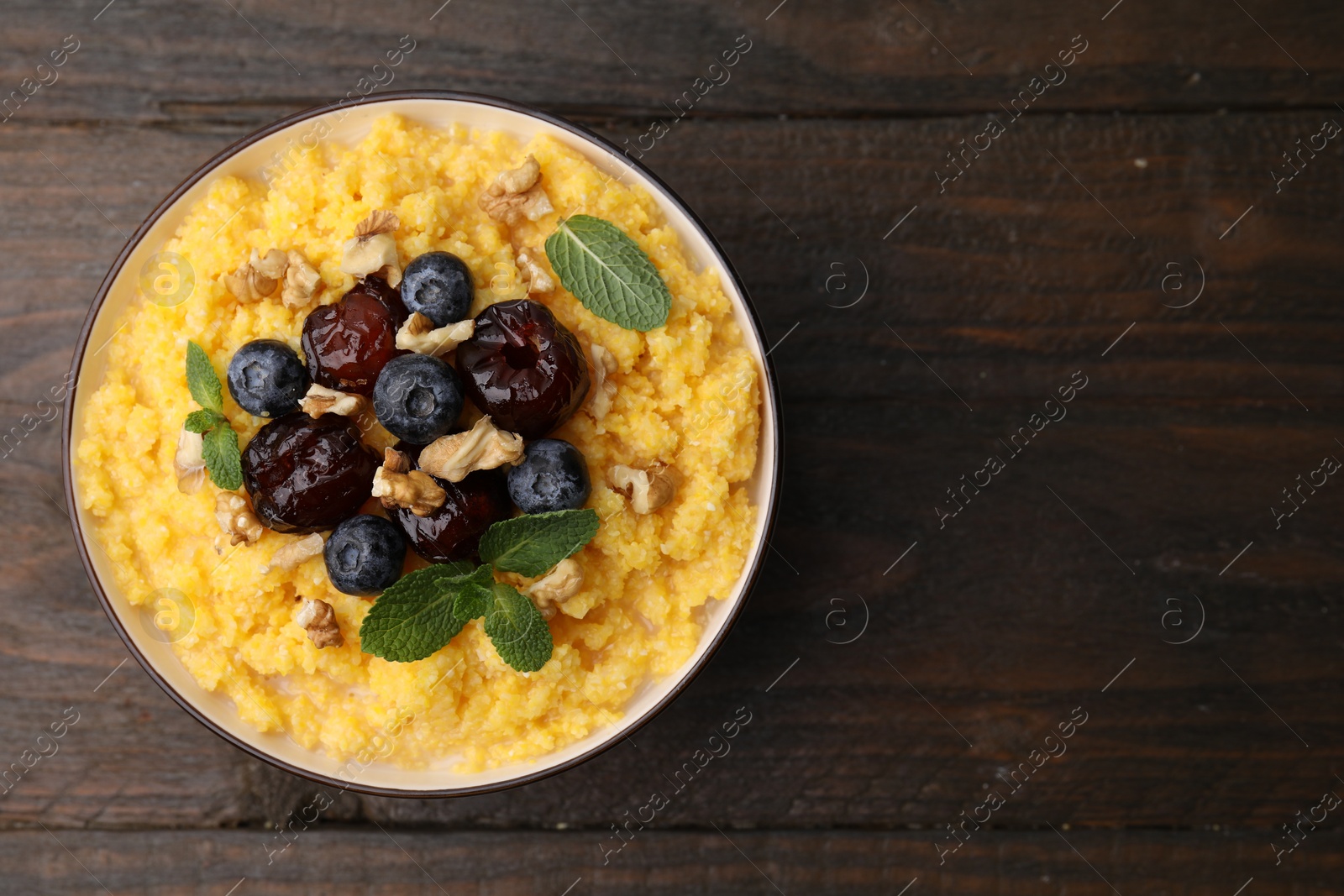 Photo of Tasty cornmeal with blueberries, dates, walnuts and mint in bowl on wooden table, top view. Space for text