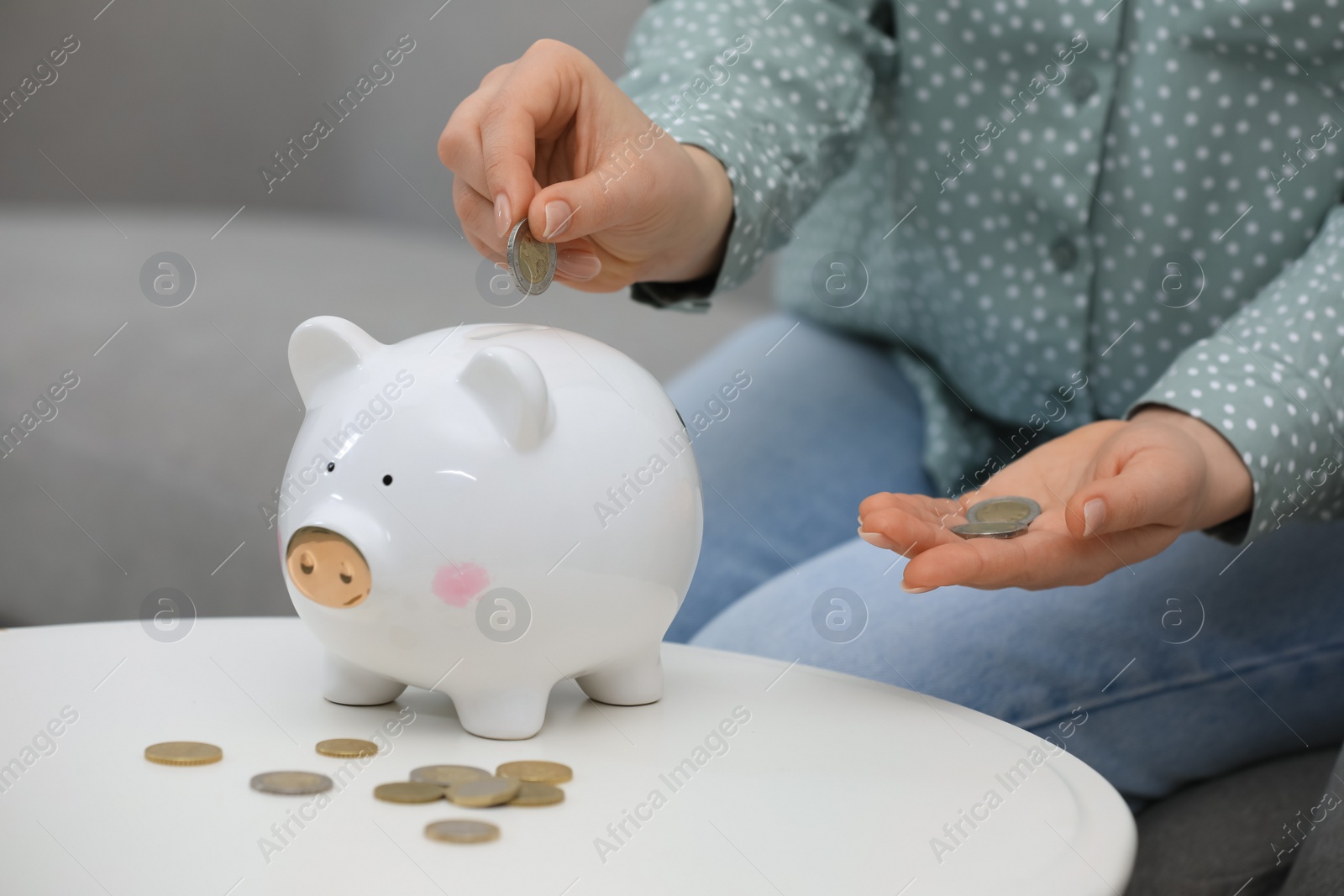 Photo of Young woman putting coin into piggy bank at table indoors, closeup