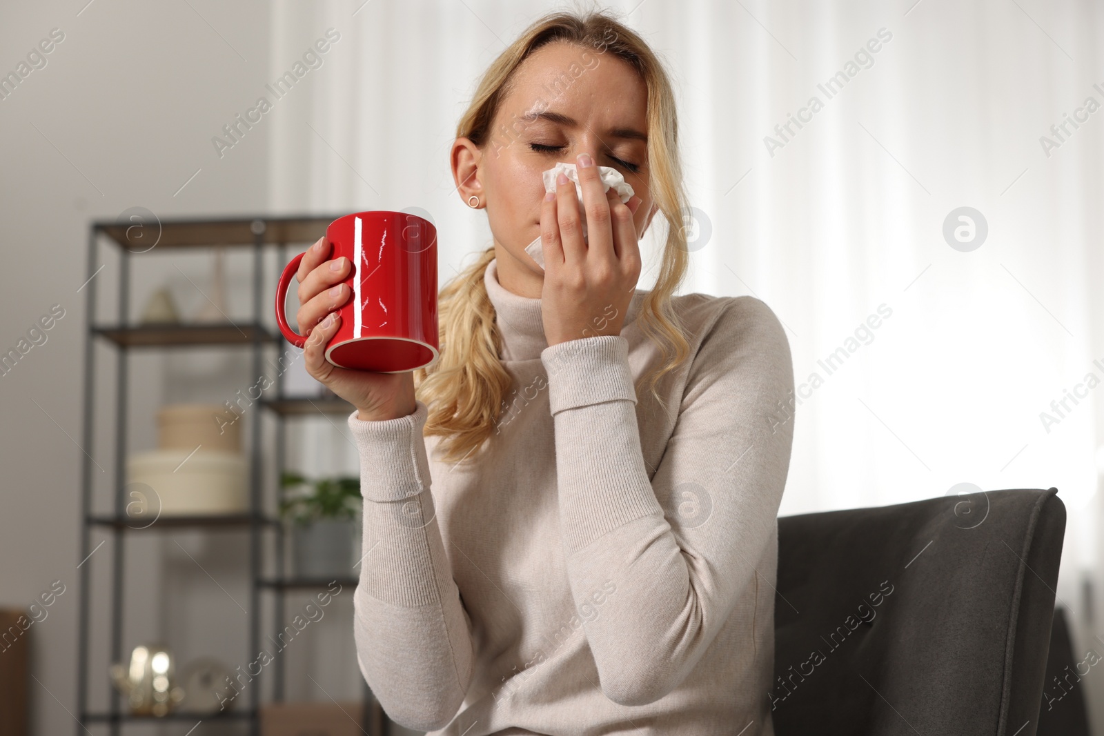 Photo of Sick woman with cup of drink blowing nose in tissue on armchair at home. Cold symptoms