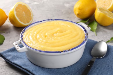 Delicious lemon curd in bowl, fresh citrus fruits, green leaves and spoon on grey table