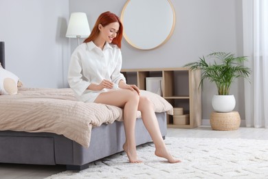 Photo of Beautiful young woman applying body cream onto legs in bedroom, space for text