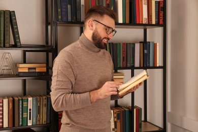 Young man reading book near shelves in home library