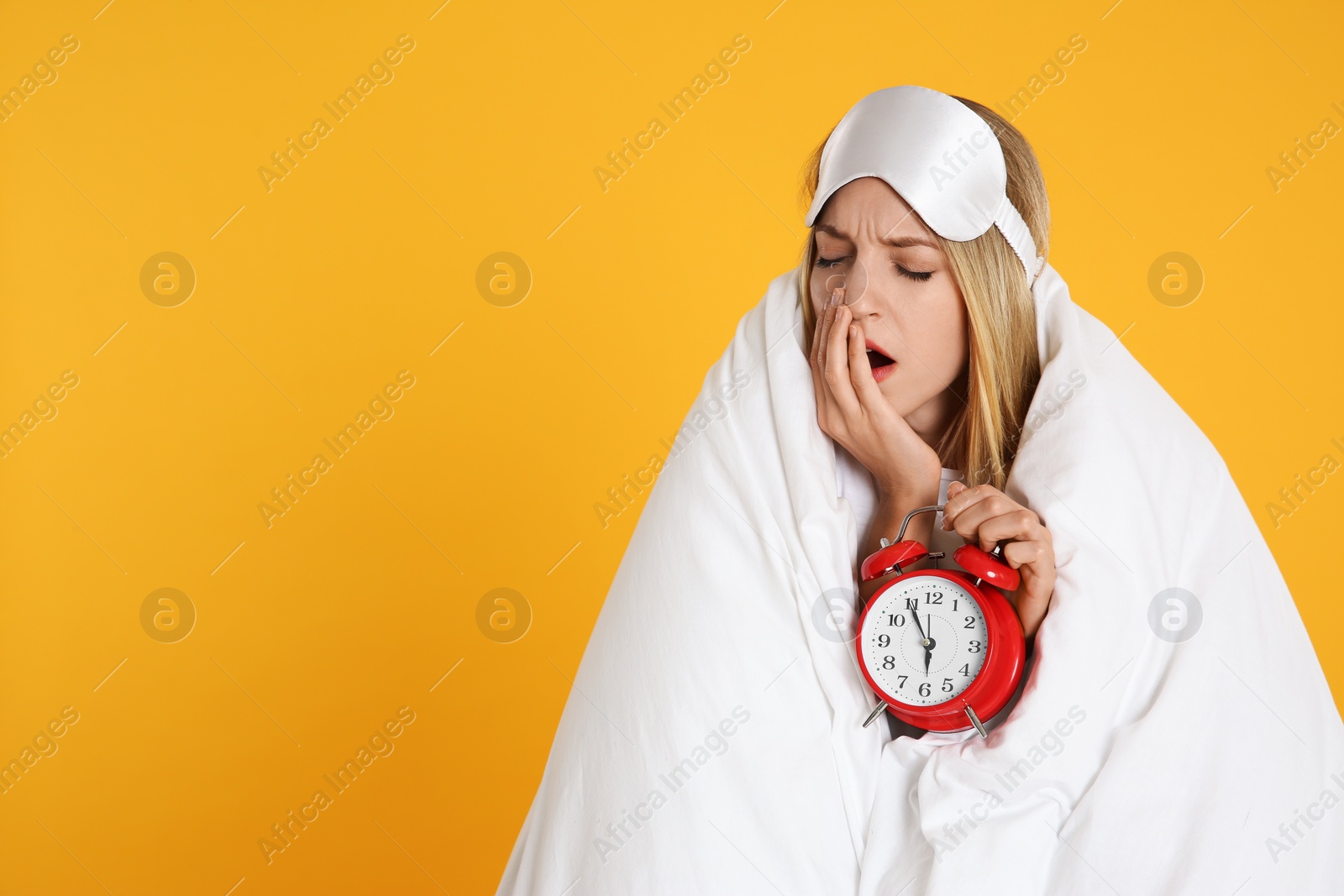 Photo of Young woman in sleeping mask wrapped with blanket holding alarm clock on yellow background. Space for text