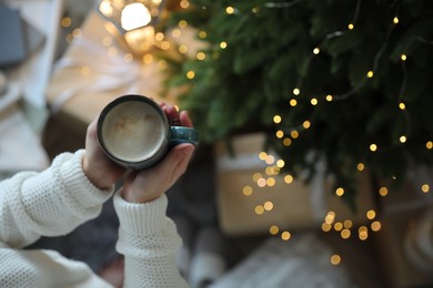 Photo of Woman holding cup of cocoa near Christmas tree indoors, top view