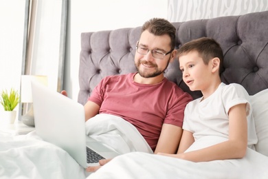 Little boy and his dad using laptop in bedroom