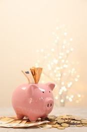 Piggy bank with euro banknotes and coins on grey table against blurred lights, space for text