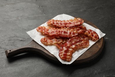 Board with fried bacon slices on dark textured table, closeup