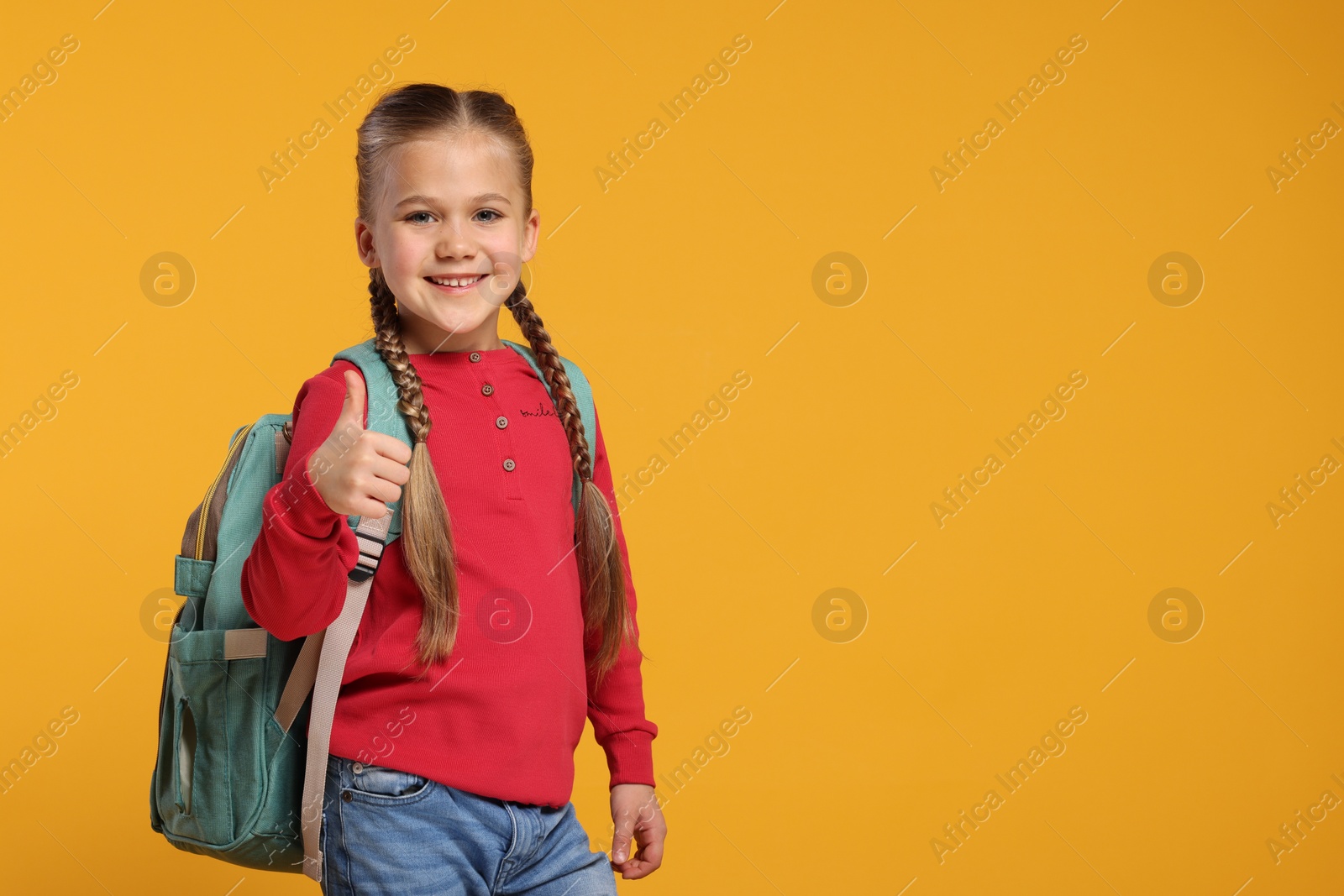 Photo of Happy schoolgirl with backpack showing thumb up gesture on orange background, space for text