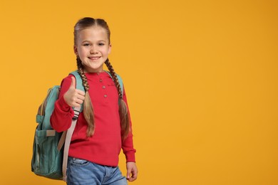 Happy schoolgirl with backpack showing thumb up gesture on orange background, space for text