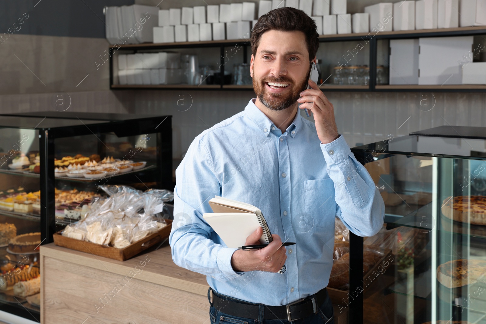 Photo of Happy business owner with notebook and pen talking on phone in bakery shop