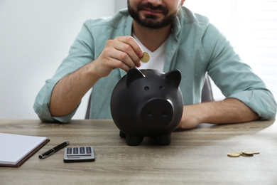 Photo of Man putting coin into piggy bank at wooden table indoors, closeup