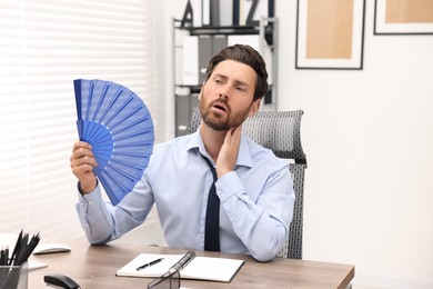 Bearded businessman waving blue hand fan to cool himself at table in office