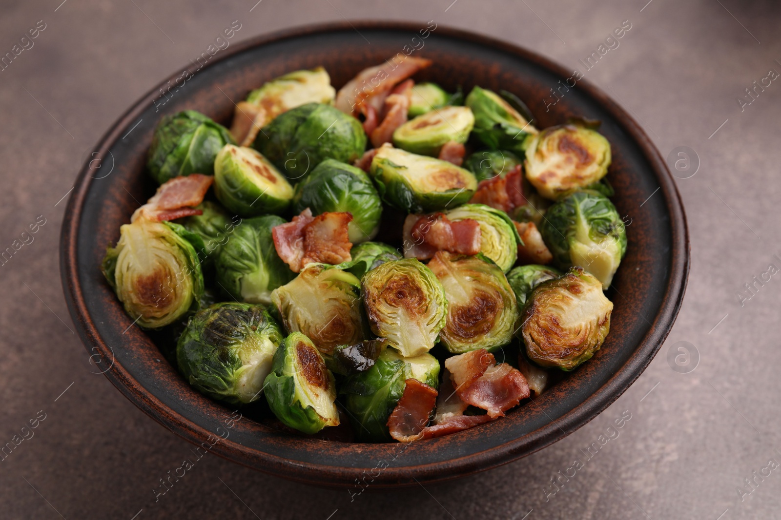 Photo of Delicious roasted Brussels sprouts and bacon in bowl on brown table, closeup