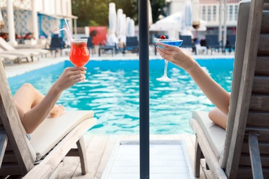 Happy young friends with fresh summer cocktails relaxing on sunbeds near swimming pool