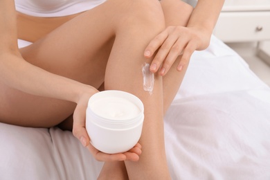 Photo of Young woman applying cream on her legs on bed, closeup. Beauty and body care