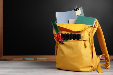 Photo of Backpack with different school stationery on white wooden table near blackboard, space for text
