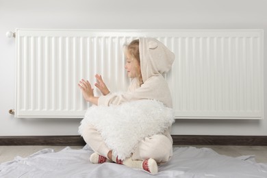 Photo of Little girl warming hands near heating radiator indoors