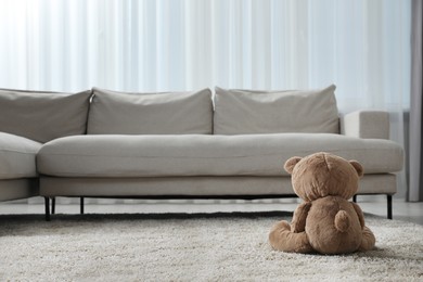 Photo of Cute lonely teddy bear on floor near sofa in room, back view. Space for text