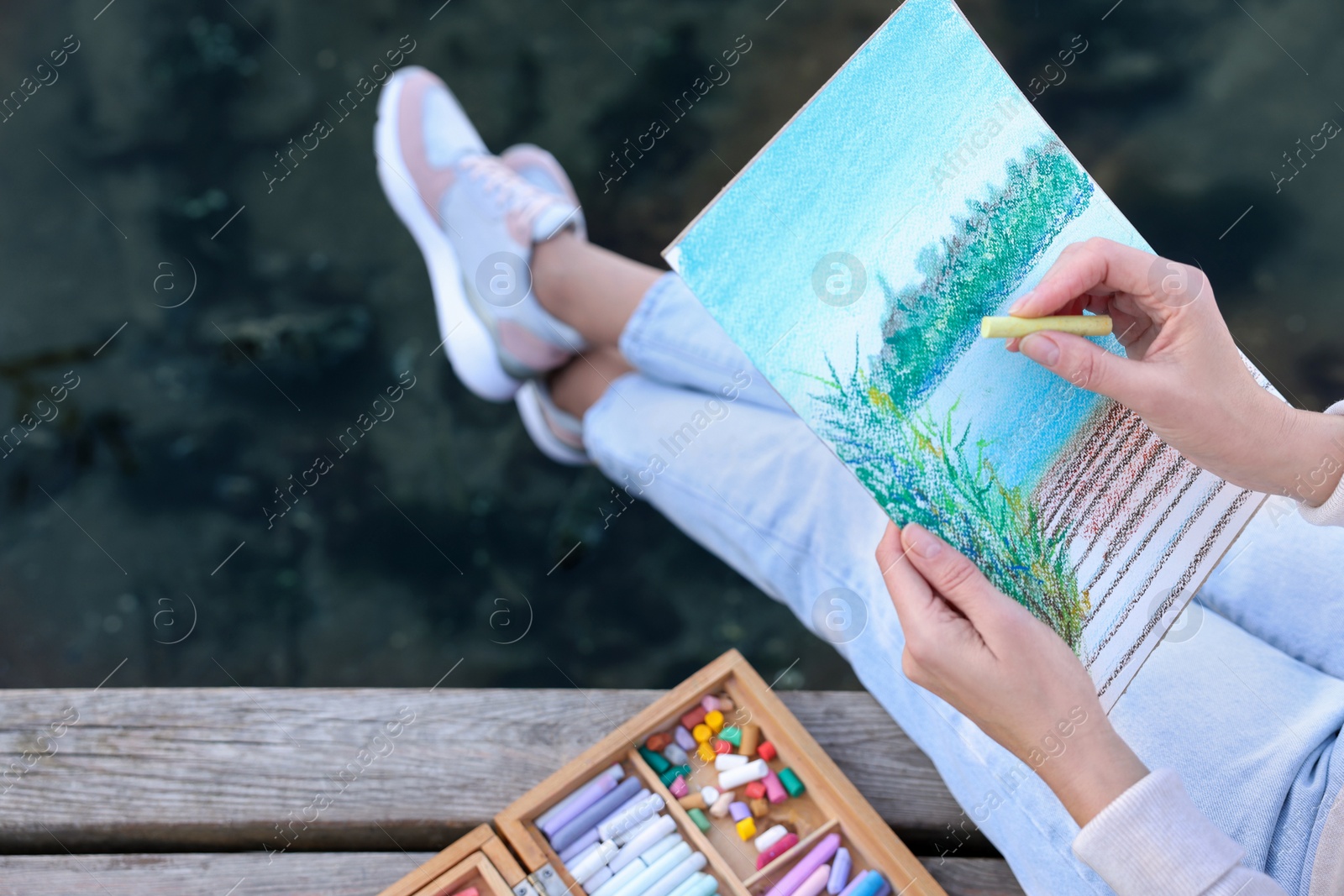 Photo of Woman drawing with soft pastels on wooden pier near river, closeup