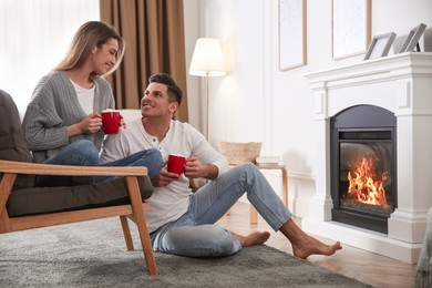 Photo of Happy couple with cups of hot drink resting near fireplace at home
