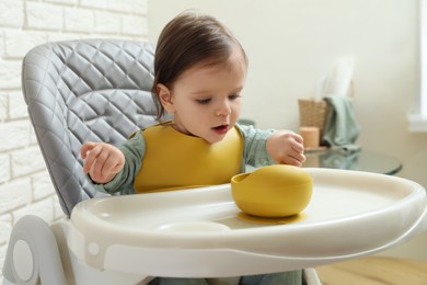 Cute little baby with bowl sitting in high chair indoors