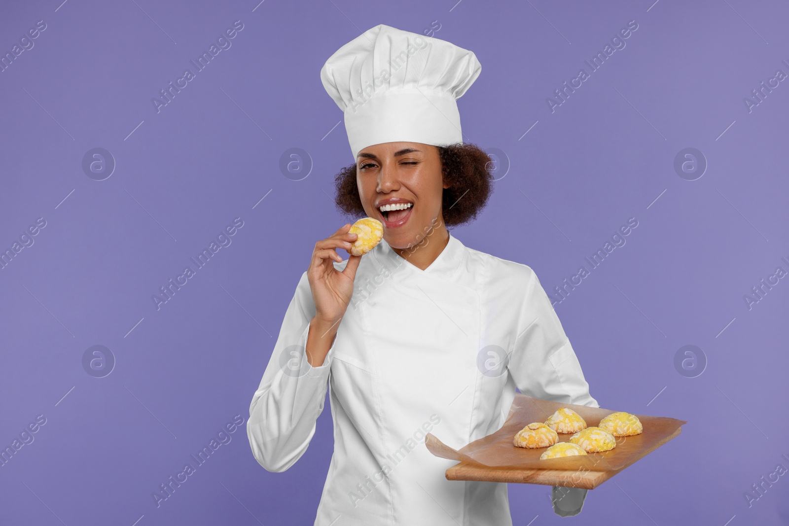 Photo of Happy female chef in uniform holding board with cookies on purple background