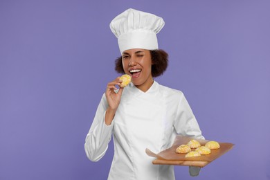 Happy female chef in uniform holding board with cookies on purple background