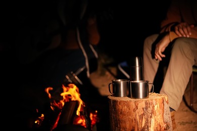 People near bonfire outdoors at night, focus on stump with metal mugs and thermos. Camping season