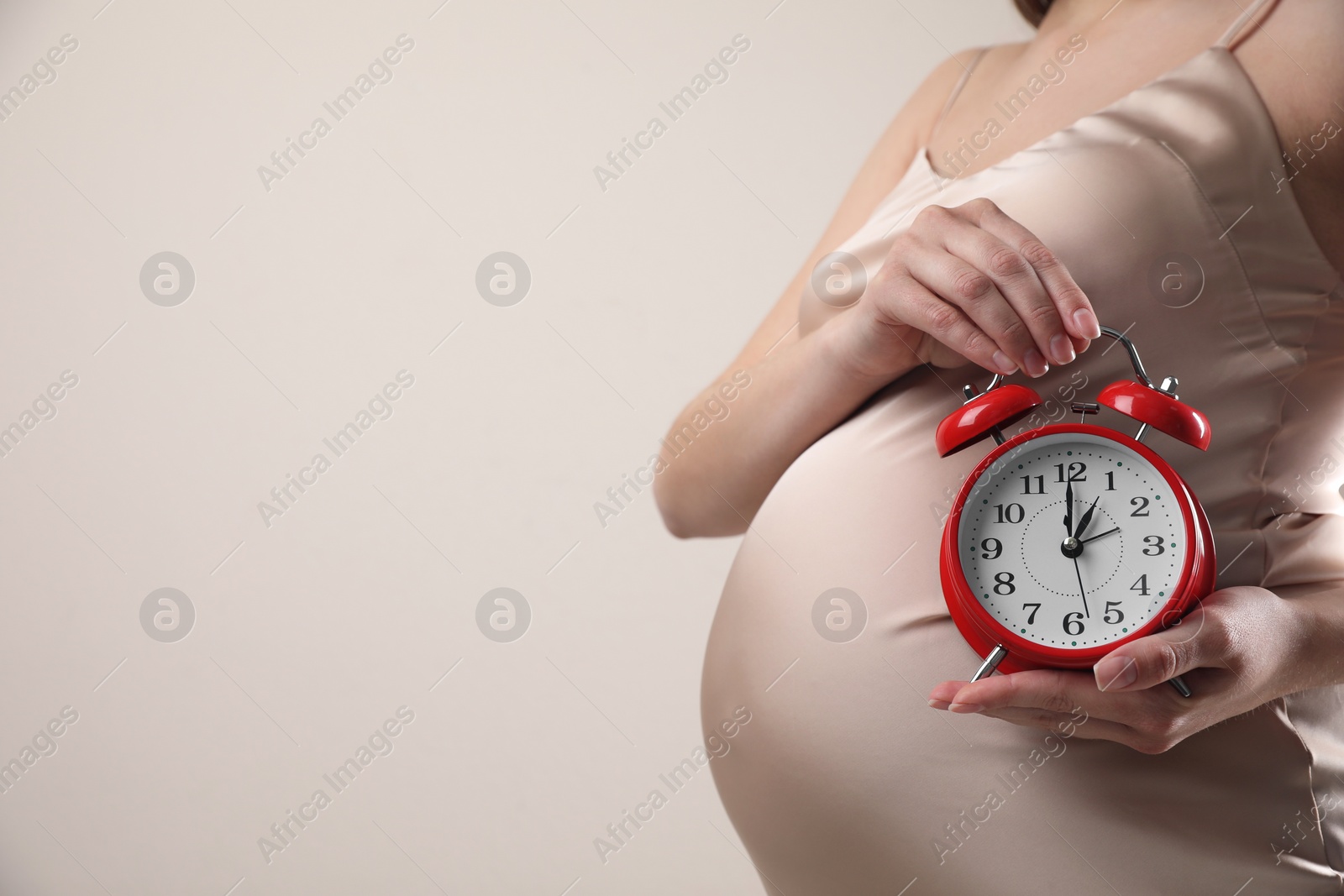 Photo of Young pregnant woman holding alarm clock near her belly on beige background, closeup and space for text. Time to give birth