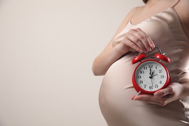Young pregnant woman holding alarm clock near her belly on beige background, closeup and space for text. Time to give birth