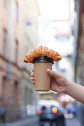 Photo of Woman holding tasty croissant and cup of coffee on city street, closeup