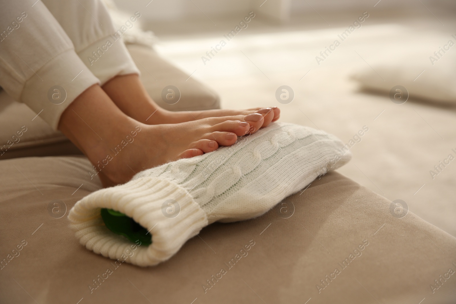 Photo of Woman warming feet with hot water bottle on sofa, closeup