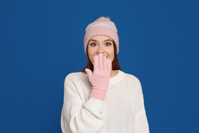 Photo of Emotional woman wearing warm sweater, gloves and hat on blue background. Winter season