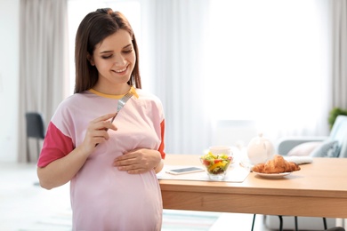Photo of Young pregnant woman eating vegetable salad near table in kitchen