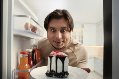 Photo of Happy overweight man taking cake out of refrigerator in kitchen, view from inside