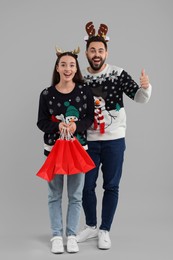 Photo of Happy young couple in Christmas sweaters and reindeer headbands with shopping bags on grey background