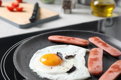 Egg and sausages in frying pan on stove indoors, closeup