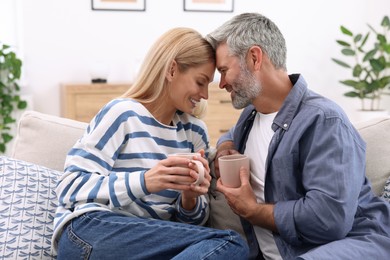 Happy affectionate couple with cups of hot drinks on sofa at home. Romantic date