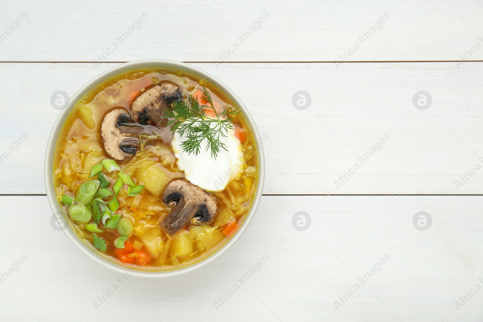 Photo of Bowl of delicious sauerkraut soup with mushrooms, green onion, dill and sour cream on white wooden table, top view. Space for text