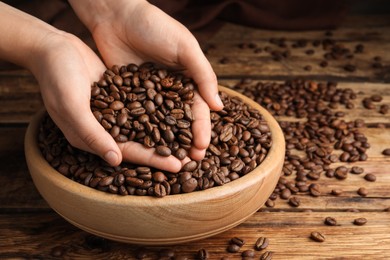Photo of Woman taking pile of roasted coffee beans from bowl at wooden table, closeup