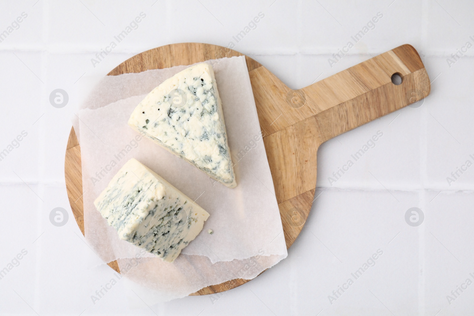 Photo of Wooden cutting board with blue cheese on white tiled table, top view