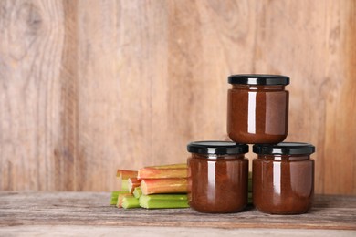 Photo of Jars of tasty rhubarb jam and stalks on wooden table, space for text