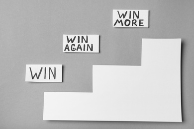 Photo of Cards with words WIN, WIN AGAIN, WIN MORE and paper cutout stairs on grey background, top view. Victory concept