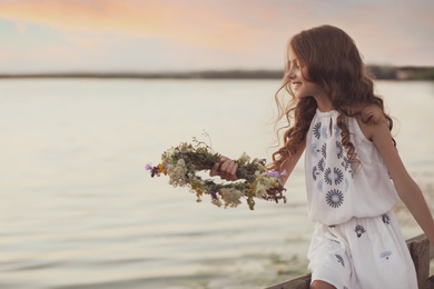 Cute little girl holding wreath made of beautiful flowers near river in evening